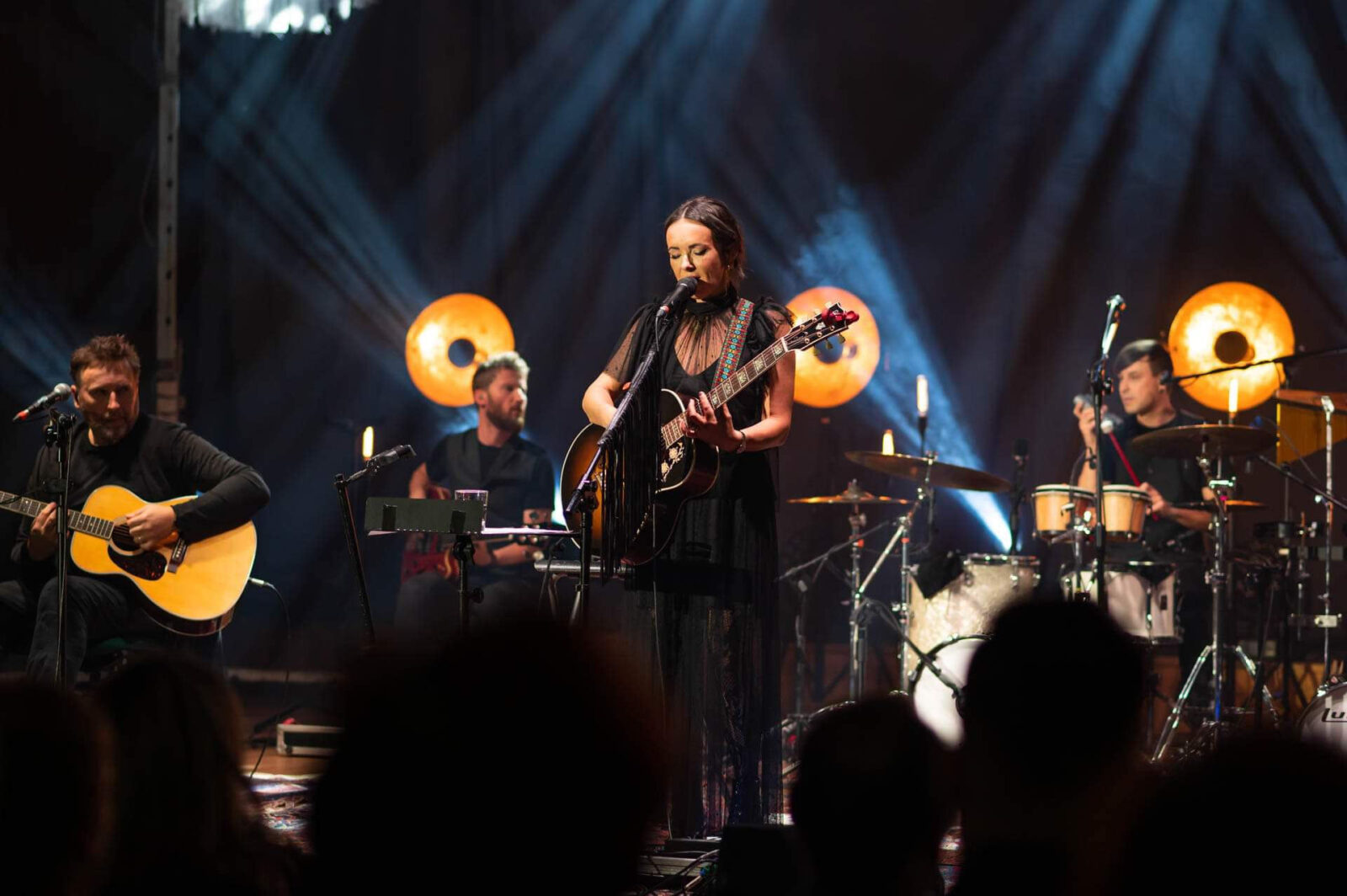 Rockowy zespół z polską wokalistką na ciemnej scenie oświetlonej punktowym światłem